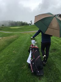 Rear view of golfer pulling golf bag on grass with umbrella against sky during rain