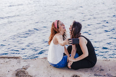 Lesbian couple taking selfie while sitting by sea