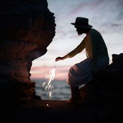 Full length of woman holding sparkle while sitting on rock against sky during sunset