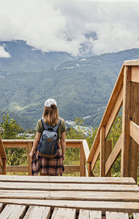 Rear view of young blonde woman  with backpack looking at view of mountains on wooden viewpoint