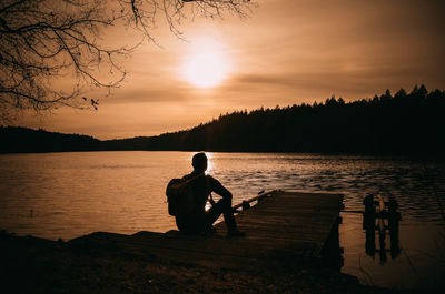 Silhouette man sitting by lake against sky during sunset