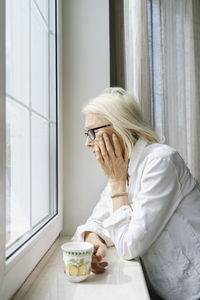 Depressed senior woman with cup leaning on window sill at home