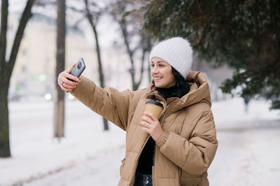 Cute girl laughing and making a video call outside in winter. hold a disposable cup of coffee