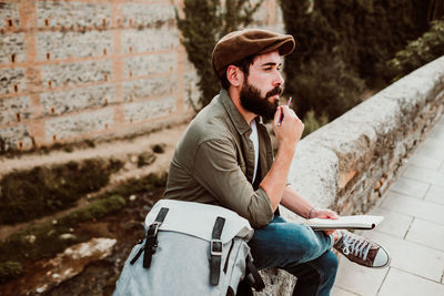 Man with backpack holding pencil and book while sitting outdoors