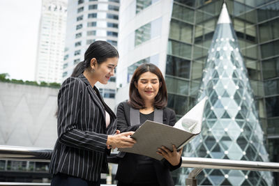 Smiling businesswomen looking at folder while standing against buildings in city