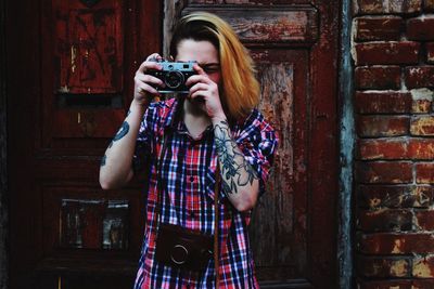 Woman with tattoo photographing through camera against brick wall