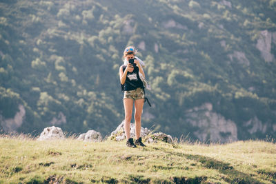 Full length of female hiker photographing while standing on field against mountain