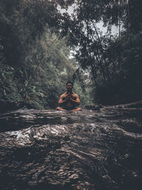 Young man meditating while sitting in stream against trees