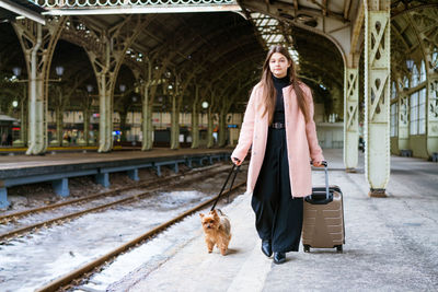 Beautiful young casual tourist woman with dog and suitcase waiting for train person