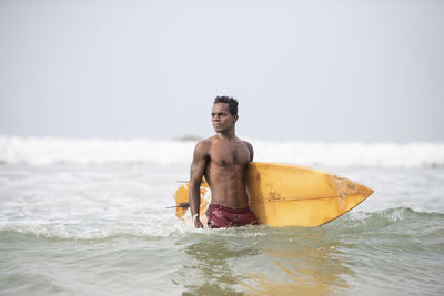 Shirtless man with surfboard standing in sea against clear sky