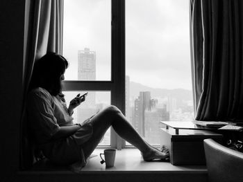 Side view of woman using phone while sitting by window at home