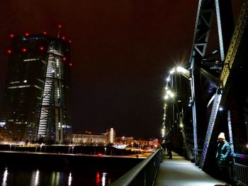 Illuminated bridge over river by buildings against sky at night