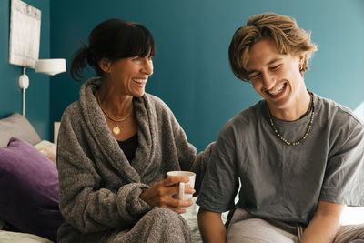 Woman holding mug while taking to happy son sitting on bed at home