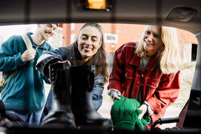 Smiling young man looking at friends loading shoes and sweater in car trunk