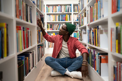 Young african american guy sits on floor in library between bookshelves, student searching for book