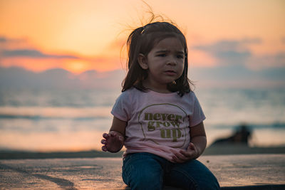 Cute upset girl sitting on retaining wall against sky during sunset