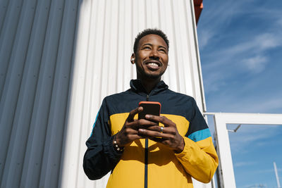 Smiling sportsman with mobile phone looking away while standing against wall