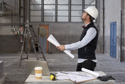 Male architect holding blueprint looking up while standing in building
