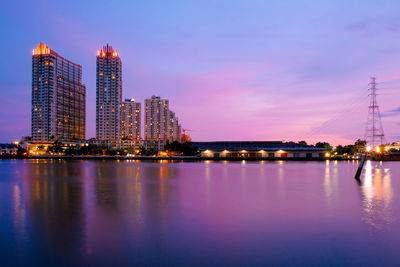 Illuminated city by river against sky at dusk