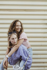 Teenage girl carrying female friend piggyback against metal wall