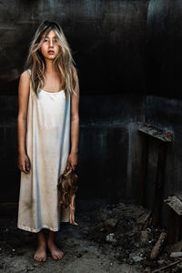 A teenage girl stands in a burned-out house with a doll in her hands