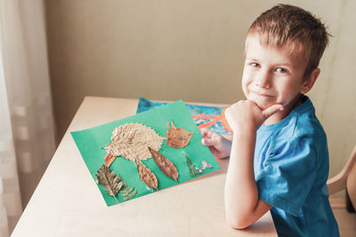 Cute boy demonstrate applique made of autumn leaves, happy child sitting by desk