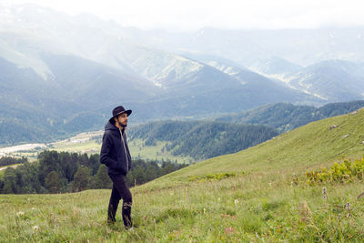 Man with a beard shepherd standing in the mountains in a black raincoat in the rain