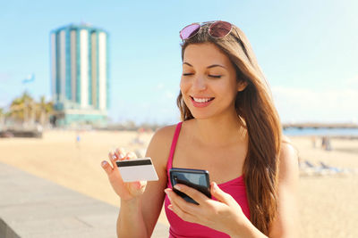 Young woman holding credit card while using mobile phone at beach