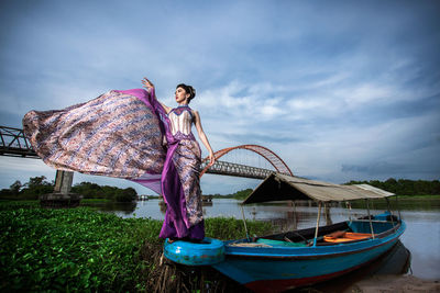 Low angle view of young model wearing dress while standing on boat in river against cloudy sky