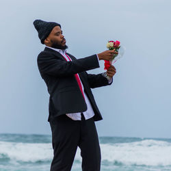 Man in formalwear holding bouquet while standing at beach against sky