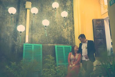 Illuminated lanterns hanging by couple looking at each other against building