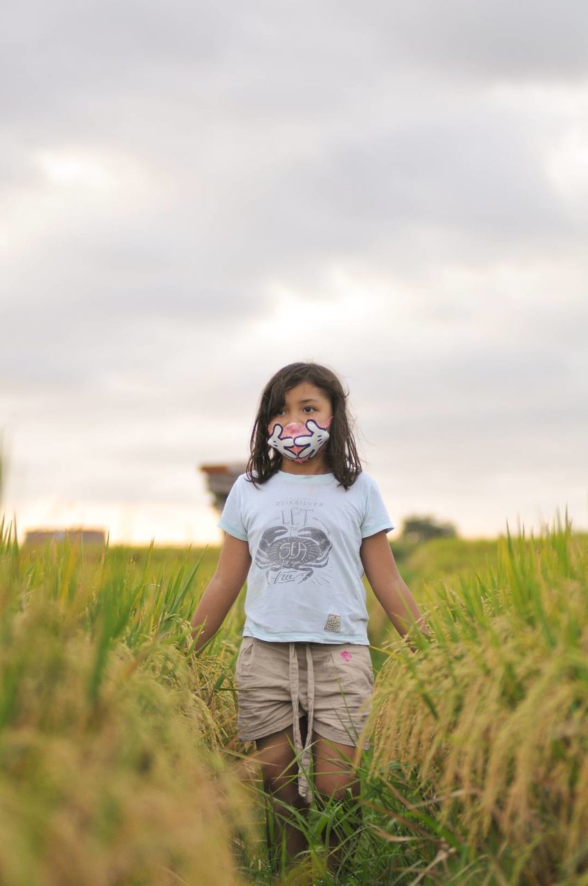 one person, grass, sky, plant, nature, field, land, women, child, landscape, front view, childhood, rural scene, cloud, portrait, casual clothing, smiling, standing, long hair, emotion, agriculture, adult, environment, happiness, hairstyle, three quarter length, outdoors, female, looking at camera, leisure activity, day, full length, copy space, summer, young adult, meadow, plain, person, lifestyles, brown hair, beauty in nature, looking