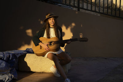 Young woman wearing hat playing guitar while sitting outdoors