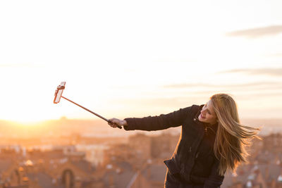Woman with arms raised standing in city against sky during sunset