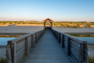 Empty footbridge leading towards water against clear sky