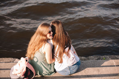 Young woman whispering into her girlfriend’s ear sitting by river