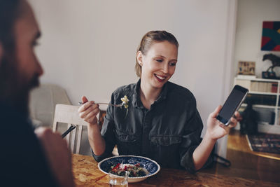 Happy woman showing mobile phone to man while eating pasta at home
