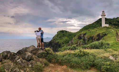 People standing on rock by lighthouse against sea and sky