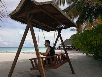 Man sitting on porch swing at beach against sky