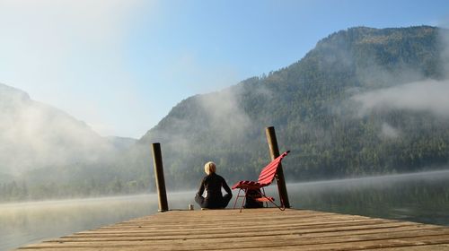 Rear view of woman sitting on pier at lake against mountain