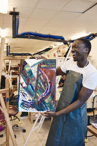 Cheerful young man showing painting while standing in art class