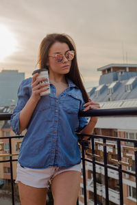 Young woman wearing sunglasses standing on balcony during sunset