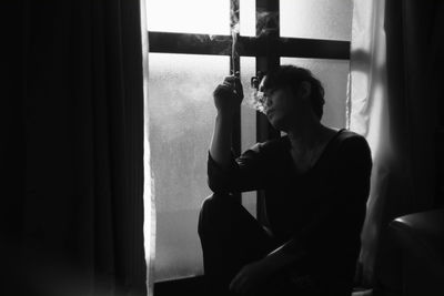 Thoughtful young man smoking cigarette while sitting by window at home