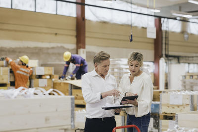 Businesswoman and businessman discussing over digital tablet while workers working in background at warehouse