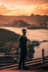 Full length of young man standing by railing against sky during sunset