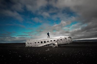 Silhouette man standing on abandoned airplane at beach against cloudy sky at night