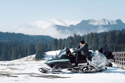 Man sitting on snowmobile in front of snowcapped mountains against sky