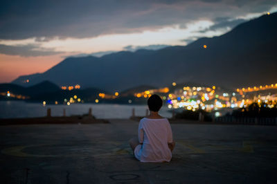 Rear view of woman looking at illuminated city by sea against sky at dusk