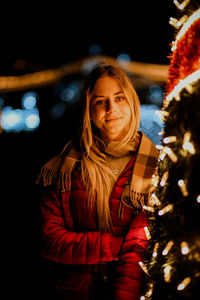 Portrait of smiling young woman standing by illuminated christmas tree at night