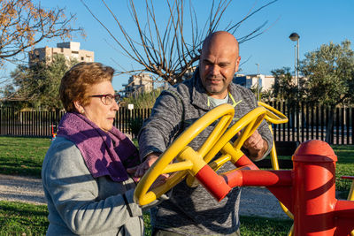 Male sports coach, helps an elderly woman to move the wheels in a bio healthy park, at sunset.